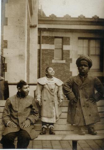 This image pictures three men, on the roof of a building (a common backdrop for Sherman's photos). At the left, seated on a bench, we see a man with his head turned to our right, wearing a knee-length jacket. Beside him another man stands on the bench, so as to draw attention to his smaller stature–he stands at about the same head-level as the seated man. The standing man is wearing an ankle-length jacket with fur trim, and his chin is raised in the air. On the far right, we see a third man, also standing on the bench and just slightly taller than the man in the middle of the picture. He wears a turban and his head is larger than those of the other two men. He also wears an ankle-length coat, and he holds his hands on his waist. The image is labeled 'Subramaino Pillay (Right) and Two Microcephalics.'