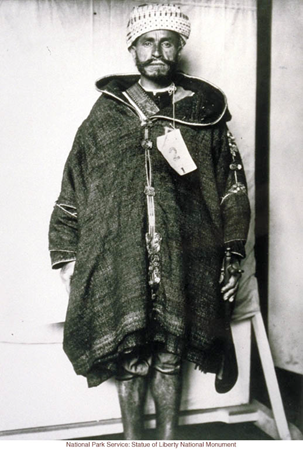 This image is labeled 'North African Immigrant,' and the picture shows a seemingly middle-aged man with a beard, a knit or woven hat, and a large hooded jacket, frayed at the bottom and closed with buttons at the front. His legs are bare. He also has a tag affixed to his jacket with the number 2 printed on it.