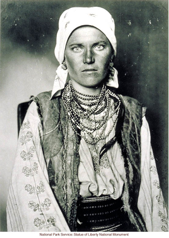 This image shows a young woman wearing a white headscarf, a vest, a blouse with floral stitching down the sleeves and front, and many beaded necklaces. She gazes directly at the camera.