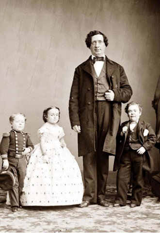 In this photo, 'General Tom Thumb' is on the far left, wearing a military outfit and holding a military hat in his hand. Beside him is Lavinia Warren, wearing an elaborate white gown. 'The Giant' wears a three-piece suit and stands beside Warren, fully twice her height. On the far right is Commodore Nutt, also wearing a three-piece suit.