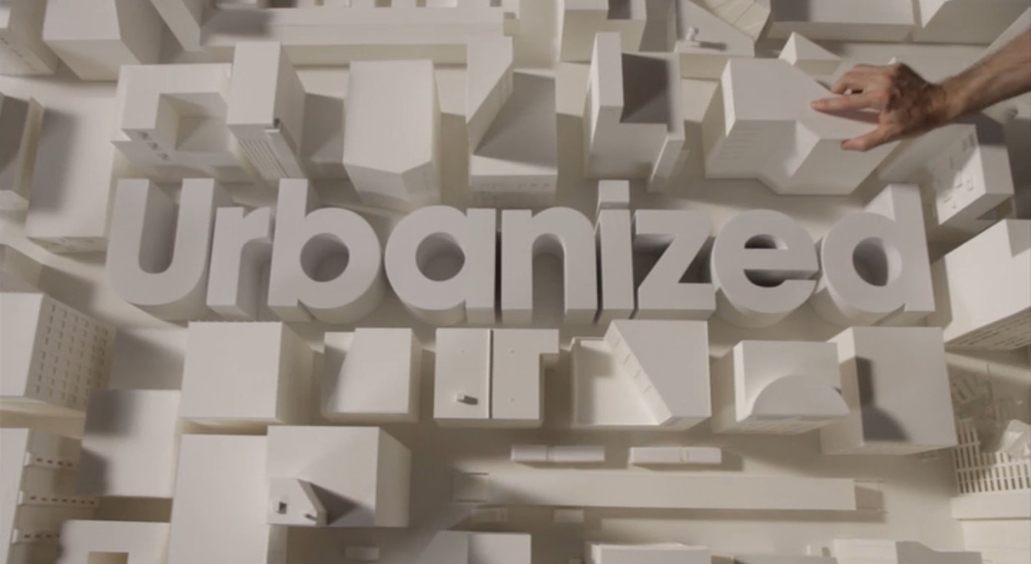 Screen capture from opening title sequence of the documentary film Urbanized. Image is a overhead shot of model buildings tightly arranged as in a city block. In the center, also as models, letters spelling 'Urbanized' have been placed. In the top right corner a human hand is placing a model building to complete the arrangement. 
